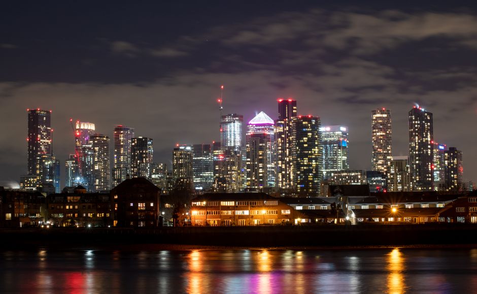 Nighttime view of the Canary Wharf skyline with illuminated modern high-rise buildings, including office towers and residential apartments, reflecting on the river below. In the foreground, a row of low-rise residential buildings with warm yellow lighting is visible along the riverbank. The image shows the urban environment of Docklands, with skyscrapers decorated with red aviation lights at their peaks, under a partly cloudy sky. This scene emphasizes the city's architecture and vibrant lights during evening hours, suitable for illustrating concepts related to house removals, furniture transport, and home relocation services offered by Removals Docklands for tight access flats in Canary Wharf.