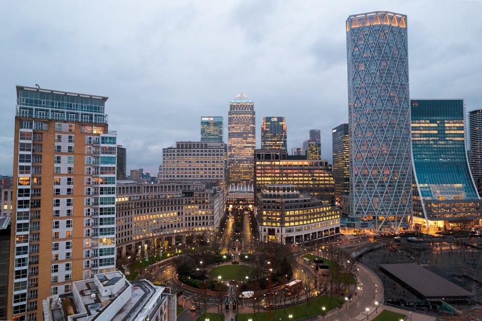 A wide-angle view of a city skyline during twilight, showcasing high-rise office buildings with illuminated windows and modern architecture. In the foreground, a residential building with a mix of glass and brick façades is visible, alongside a landscaped park with pathways and trees. Several cardboard boxes and wrapped furniture are seen inside a property, positioned near a doorway, indicating ongoing packing or home relocation preparations. A blue moving trolley is partially visible on the pavement outside, adjacent to a moving van. Overhead, overcast skies cast diffused light across the scene, emphasizing the urban environment and the logistics involved in furniture transport and house removals. The setting reflects the context of professional removals in the Docklands area, as demonstrated by the companies specializing in house moves and tight access relocations, such as Removals Docklands.