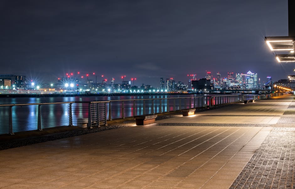 Nighttime view of the Canary Wharf skyline with illuminated modern high-rise buildings, including office towers and residential apartments, reflecting on the river below. In the foreground, a row of low-rise residential buildings with warm yellow lighting is visible along the riverbank. The image shows the urban environment of Docklands, with skyscrapers decorated with red aviation lights at their peaks, under a partly cloudy sky. This scene emphasizes the city's architecture and vibrant lights during evening hours, suitable for illustrating concepts related to house removals, furniture transport, and home relocation services offered by Removals Docklands for tight access flats in Canary Wharf.