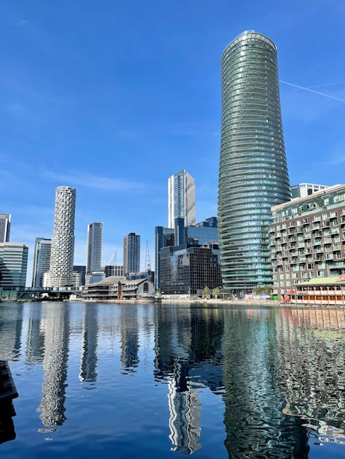 A daytime view of the Isle of Dogs waterfront showing a collection of modern high-rise buildings and office towers, including a prominent cylindrical skyscraper with glass windows reflecting the sky. The buildings are situated along the waterside, with their reflections visible in the calm water below. In the foreground, part of a dock or quay is visible, and the sky is clear with a few thin clouds. The scene captures the urban environment typical of Docklands, with natural sunlight illuminating the facades of the buildings. This imagery relates to the context of house removals or furniture transport services in the area, as facilitated by companies such as Removals Docklands, highlighting the city's skyline and potential logistical considerations for home relocation within this modern development.