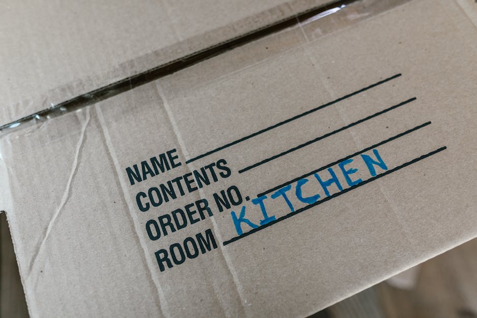 A cardboard moving box with printed fields for the owner's name, contents, order number, and room designation, which is handwritten as 'KITCHEN' in blue ink. The box is sealed with clear packing tape and is positioned on a surface inside a home, ready for a home relocation process. Nearby, there are other boxes and packing materials typically used in furniture transport and packing and moving activities, indicating an organized approach to house removals. The environment appears well-lit, highlighting the details of the box and its labels, with the scene reflecting the preparations involved in a professional removal service, such as those offered by Removals Docklands, during the loading or packing phase of a move.