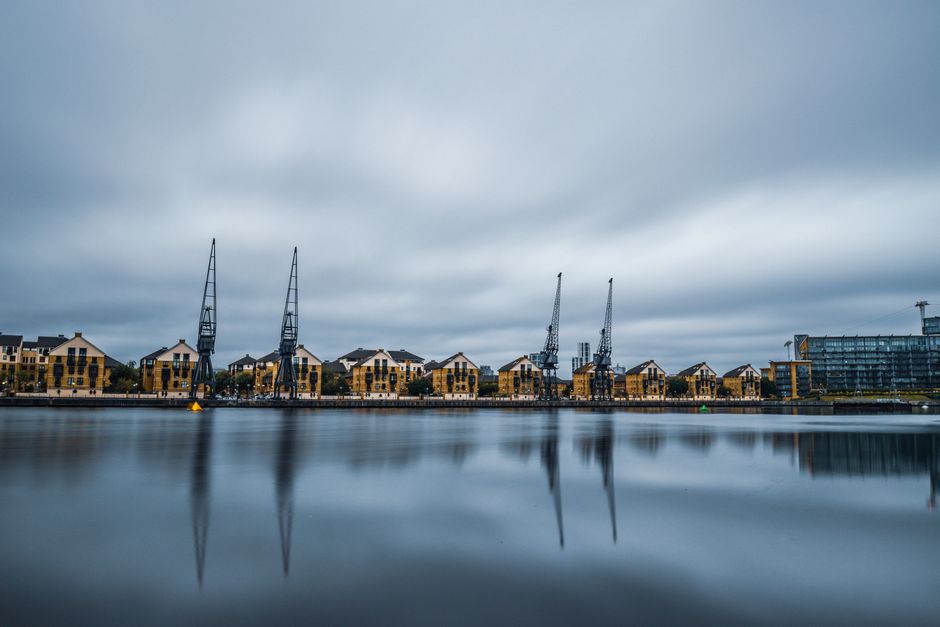 A panoramic view of a waterfront scene near Limehouse Basin in Docklands during daytime, featuring a series of modern, yellow-brick residential buildings with black window frames and pitched roofs, situated behind a calm water body that reflects their image. In front of the buildings, there are three large industrial cranes with black structural frameworks, positioned along the canal edge, used historically for shipping and now possibly signage of ongoing redevelopments. The sky overhead is overcast with grey clouds, casting diffuse lighting across the scene. On the pavement adjacent to the water, a few cardboard boxes and packing materials are visible, indicating ongoing or recent packing activities related to home relocation processes. Some furniture items, such as small chairs and tables, and protective blankets or wraps are near the boxes, suggesting careful packing before transport. A van or vehicle used for moving is not directly visible but implied by the setting. This scene, associated with Removals Docklands’ house and furniture removal services, captures the logistical environment involved in a furniture transport and packing process during a local property move near Limehouse Basin.
