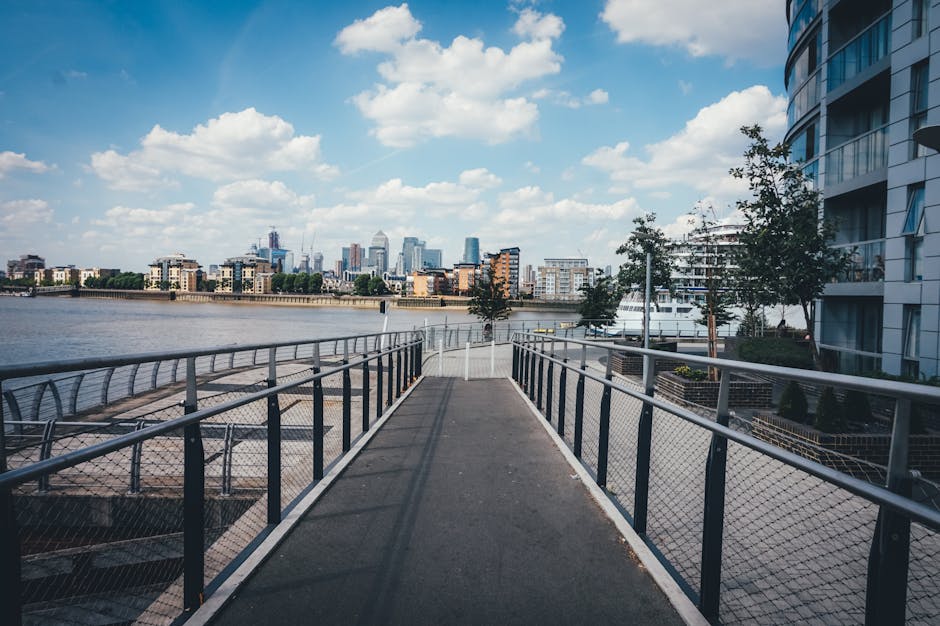 A wide, elevated outdoor walkway with metal railings and a mesh fence on both sides overlooking the River Thames in London. The pathway is made of dark asphalt and curves slightly in the distance, leading towards a cityscape of modern skyscrapers and buildings under a partly cloudy sky. On the right side, there is a multi-storey residential building with large windows and balconies, while the left side features a docking area with moored boats and yachts. The environment appears bright with natural daylight, suggesting clear weather. This setting reflects an urban riverside location suitable for home relocation or moving logistics, as part of a house removal process near Limehouse Basin, Docklands, with potential for future furniture transport or packing and moving activities. Removals Docklands specializes in managing such property moves, ensuring efficient transportation inside this urban setting.
