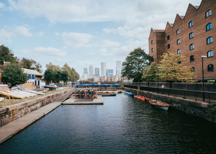 A panoramic view of a waterfront scene near Limehouse Basin in Docklands during daytime, featuring a series of modern, yellow-brick residential buildings with black window frames and pitched roofs, situated behind a calm water body that reflects their image. In front of the buildings, there are three large industrial cranes with black structural frameworks, positioned along the canal edge, used historically for shipping and now possibly signage of ongoing redevelopments. The sky overhead is overcast with grey clouds, casting diffuse lighting across the scene. On the pavement adjacent to the water, a few cardboard boxes and packing materials are visible, indicating ongoing or recent packing activities related to home relocation processes. Some furniture items, such as small chairs and tables, and protective blankets or wraps are near the boxes, suggesting careful packing before transport. A van or vehicle used for moving is not directly visible but implied by the setting. This scene, associated with Removals Docklands’ house and furniture removal services, captures the logistical environment involved in a furniture transport and packing process during a local property move near Limehouse Basin.
