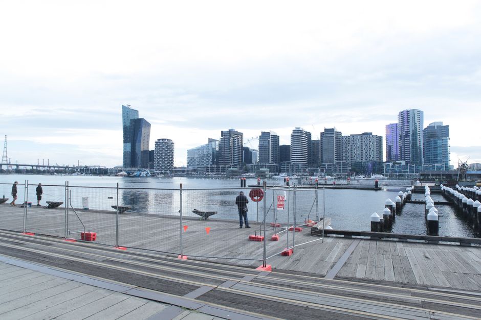 A wide view of the Royal Victoria Dock in Docklands shows modern residential buildings with yellow facades and dark roof structures lined along the waterfront. Several large industrial cranes are posi