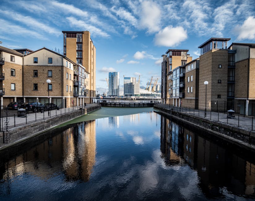 A wide view of a residential canal in Docklands with modern multi-storey apartment buildings on either side, constructed from light brown brick and featuring balconies, large windows, and flat roofs.